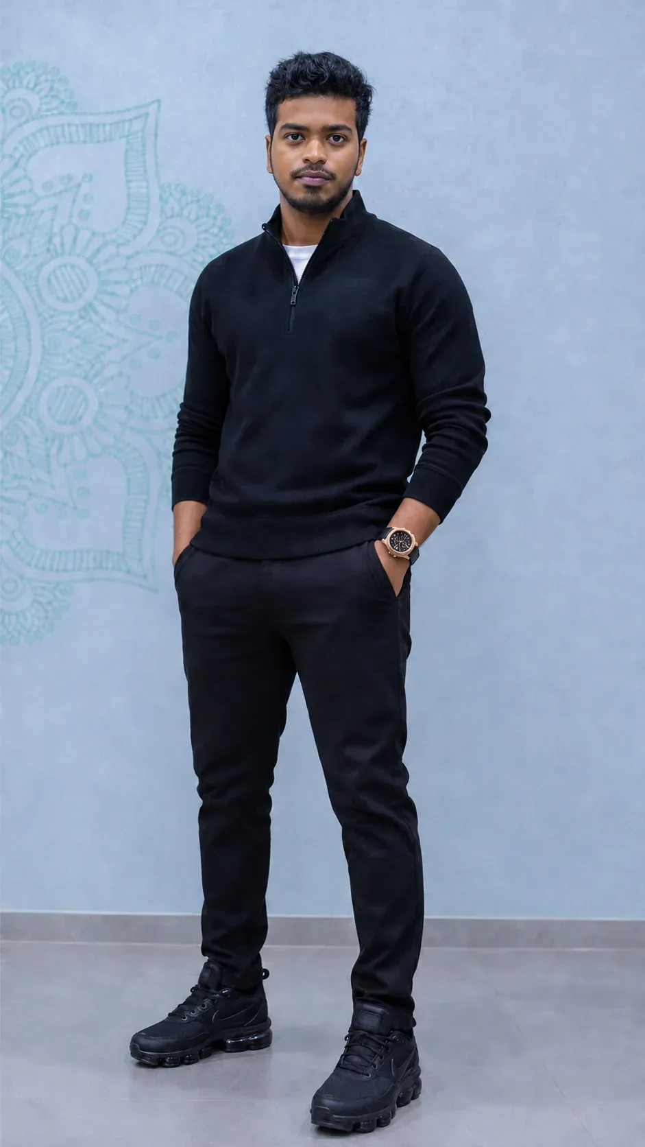 Naveen Balasubramaniam, standing in a black quarter-zip sweater against a pale blue wall with a teal mandala
